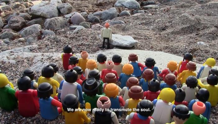 Rows of colorful toy figurines facing two people and a small boat on a rocky shore.