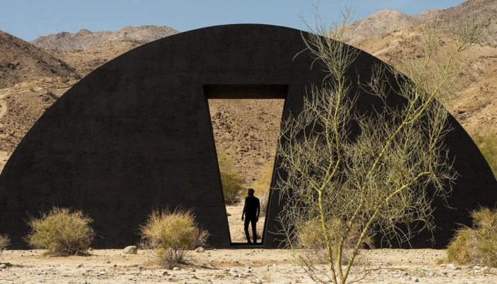 Image of Torkwase Dyson's installation at Desert X. A black semicircular form in a desert landscape. A rectangular void has been created at center and human figure stands in the space.