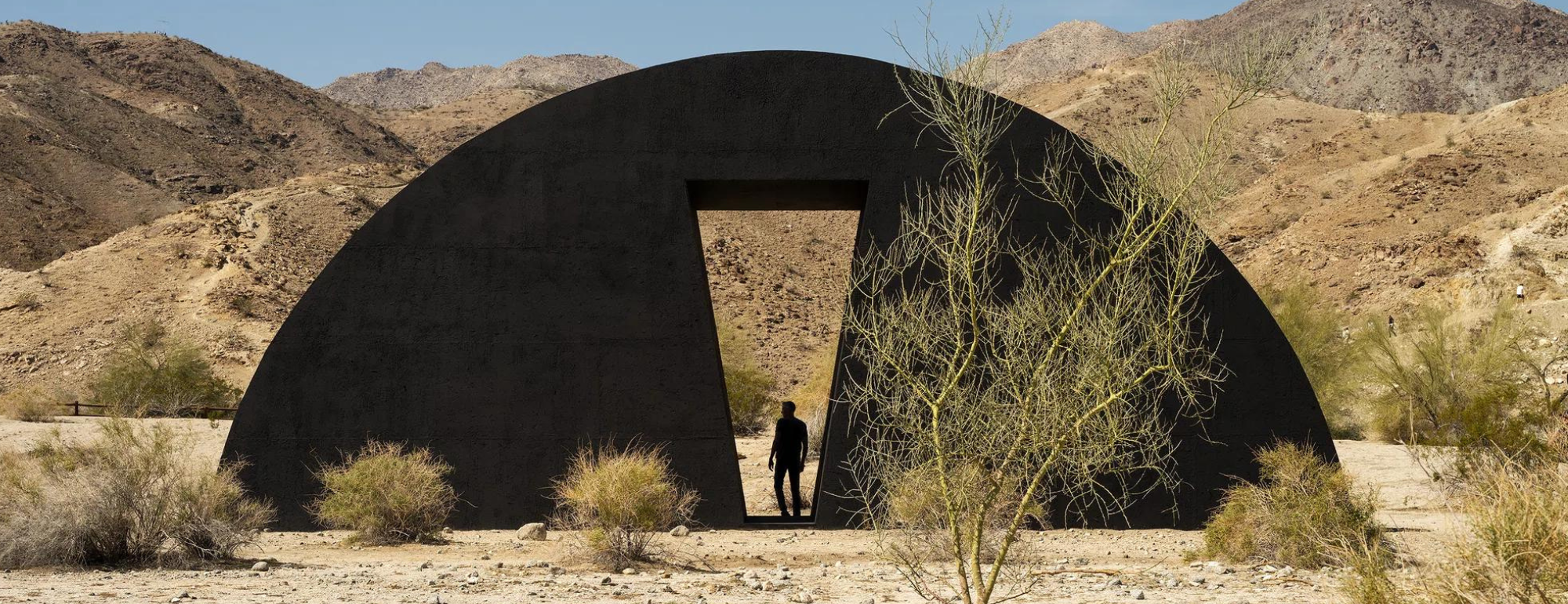 Image of Torkwase Dyson's installation at Desert X. A black semicircular form in a desert landscape. A rectangular void has been created at center and human figure stands in the space.