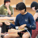 A student concentrated on weaving at a woodworking table, with classmates in the background.
