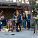 Musicians playing various instruments outdoors, surrounded by trees and buildings.